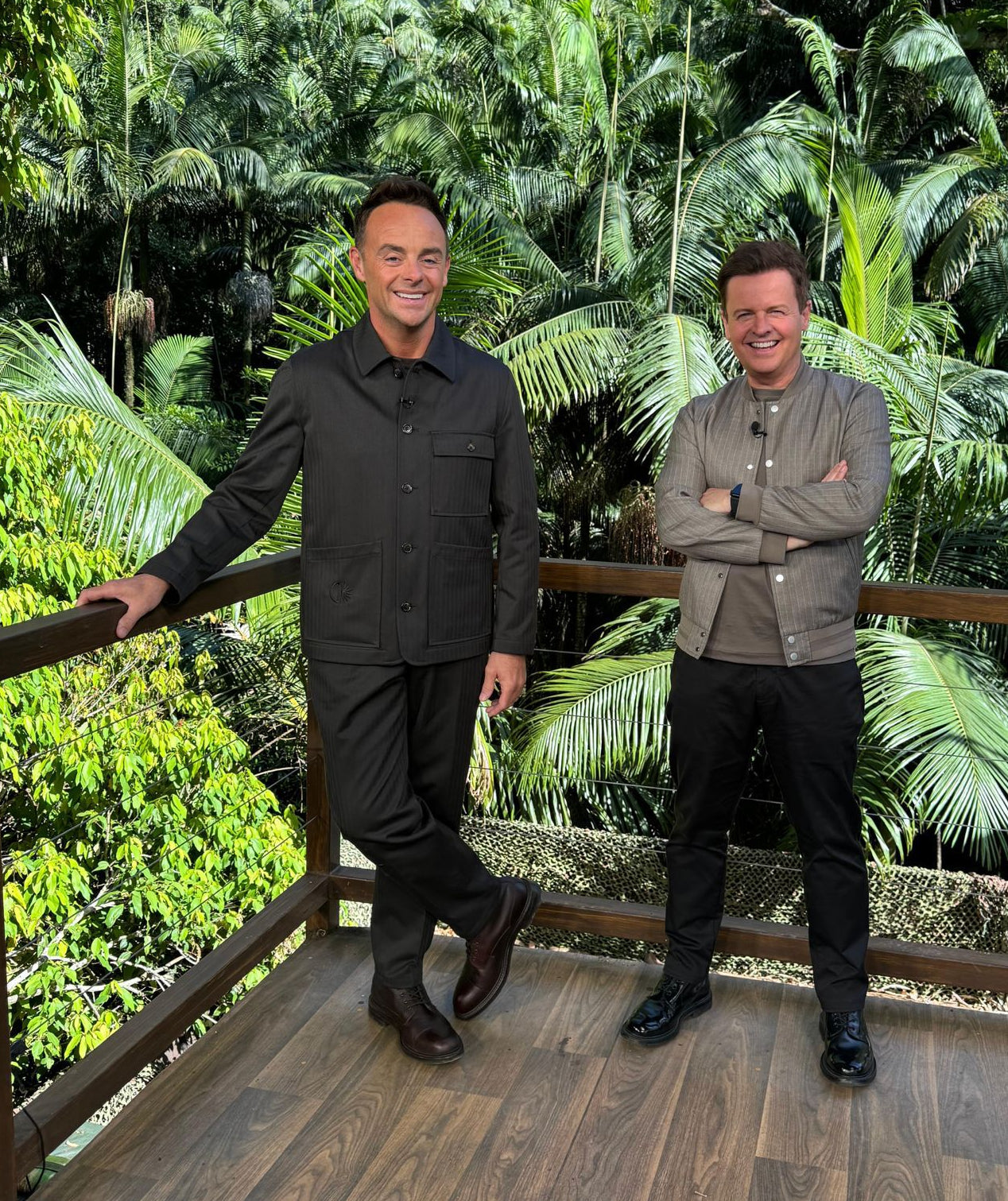 Two men standing on a wooden deck surrounded by dense tropical green foliage. The man on the left wears a black button-up shirt and black pants, leaning on a wooden railing.