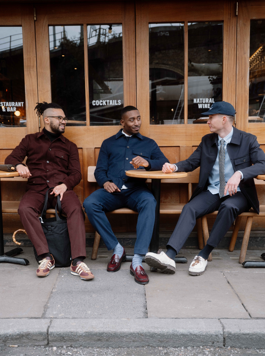 Three men in stylish menswear sit on outdoor cafe benches in front of a restaurant with wood-framed windows. The outfits include a burgundy two-piece set, a navy blue set, and a dark gray suit with a light blue shirt and tie.
