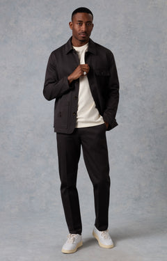Man wearing a dark jacket, white shirt, black pants, and white sneakers against a gray background