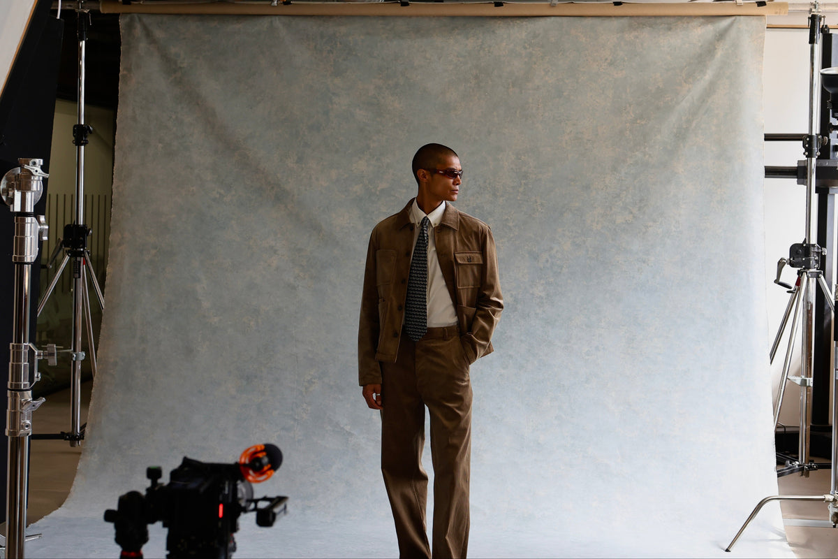 A man wearing a brown suit, white shirt, and dark tie stands in front of a textured gray studio backdrop, with photography equipment visible around him in a studio setting.
