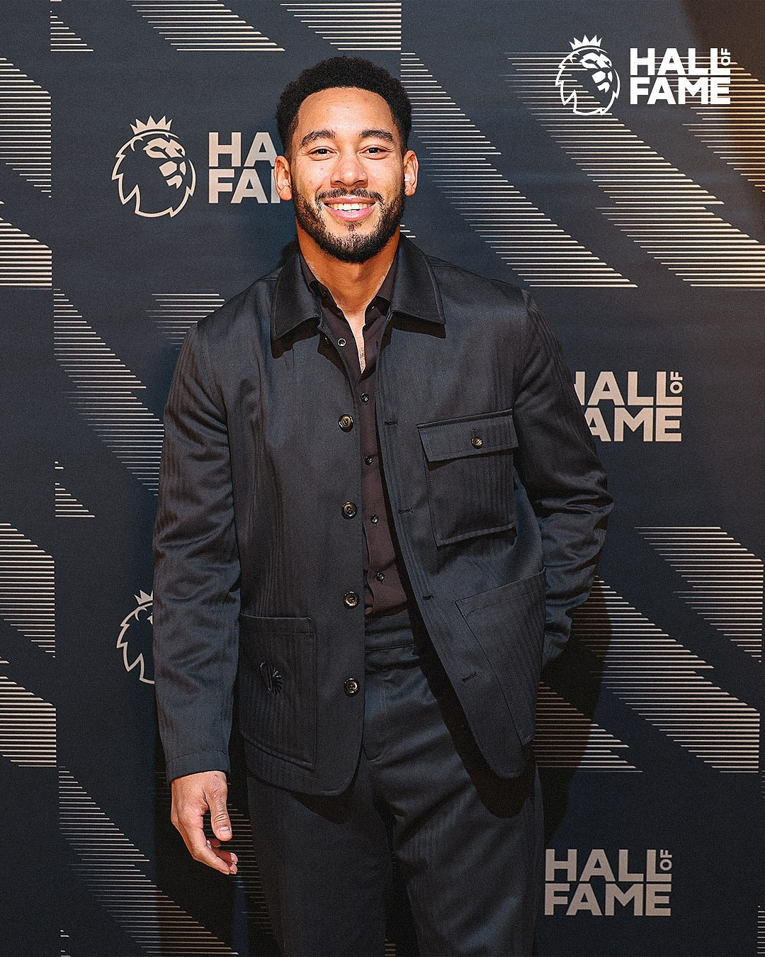 A smiling man wearing a dark charcoal pinstripe jacket and matching trousers stands in front of a dark patterned 'HALL OF FAME' backdrop with the Lions logo.