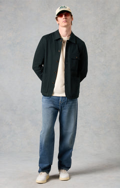 Man wearing a black jacket, white shirt, blue jeans, and a cap against a gray background