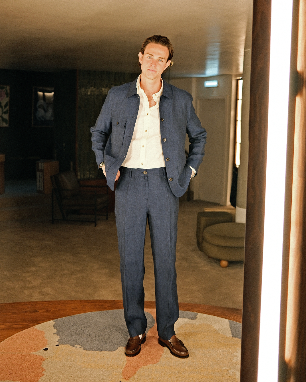 A man wearing a blue textured linen-blend suit with a white shirt and brown leather loafers, standing in a modern, dimly lit interior room.