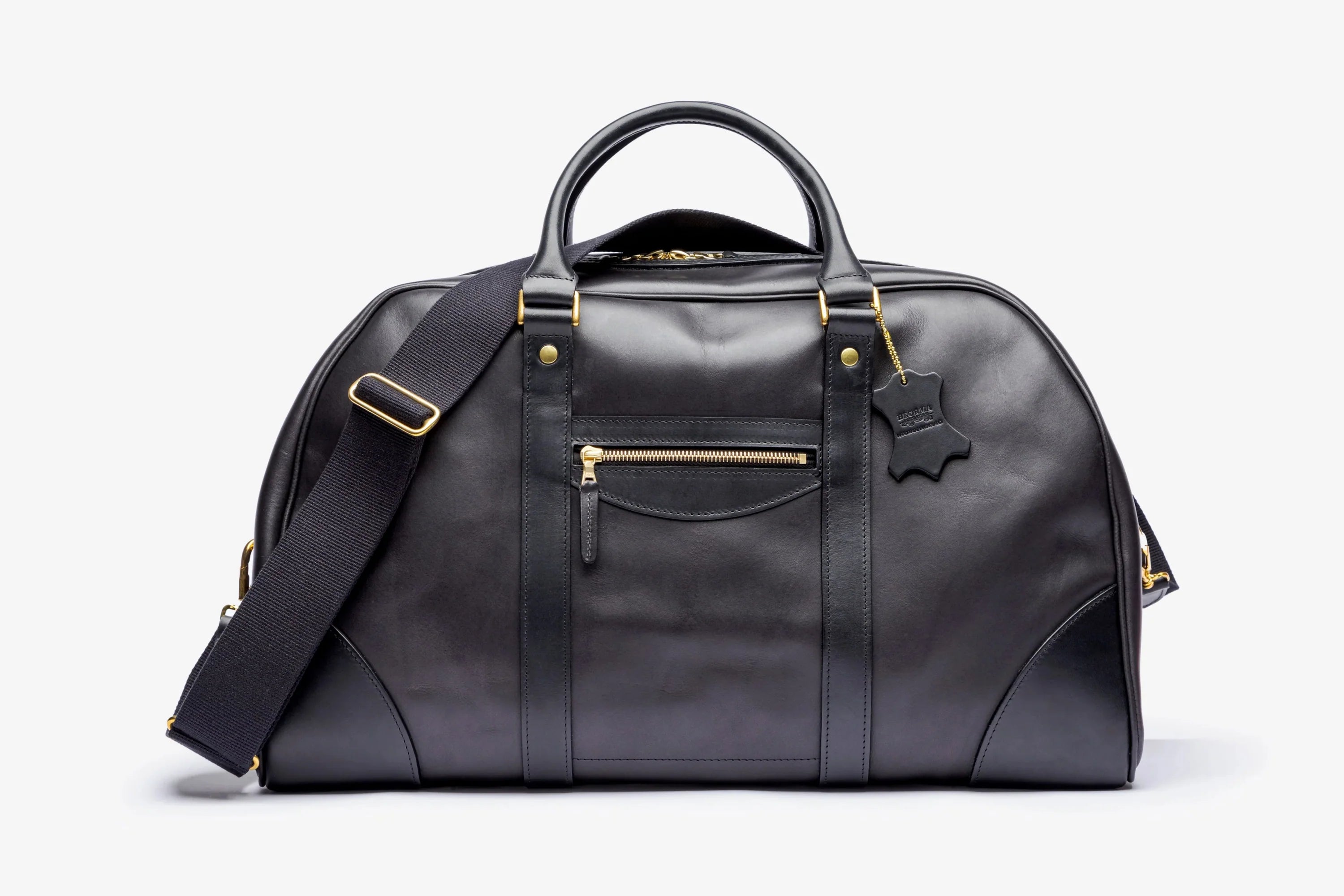 A black leather duffel bag with gold-tone hardware, featuring dual top handles, a front zippered pocket, and an adjustable shoulder strap, photographed on a clean white background.