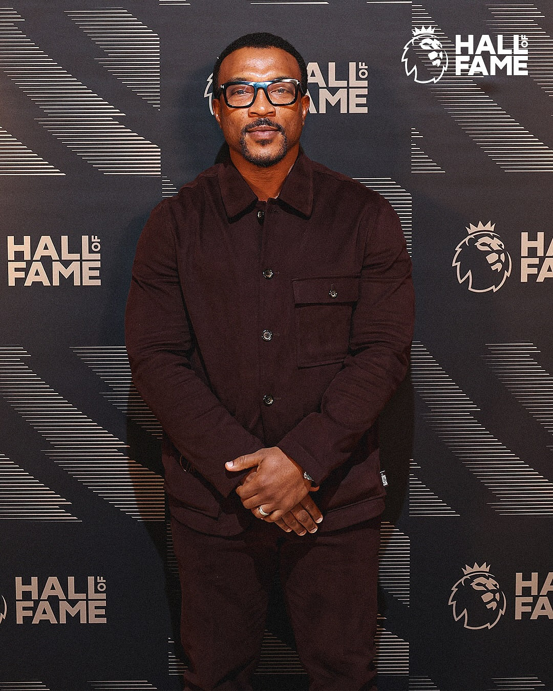 A man wearing a dark brown corduroy button-up shirt and matching trousers stands in front of a black and white 'Hall of Fame' branded backdrop.