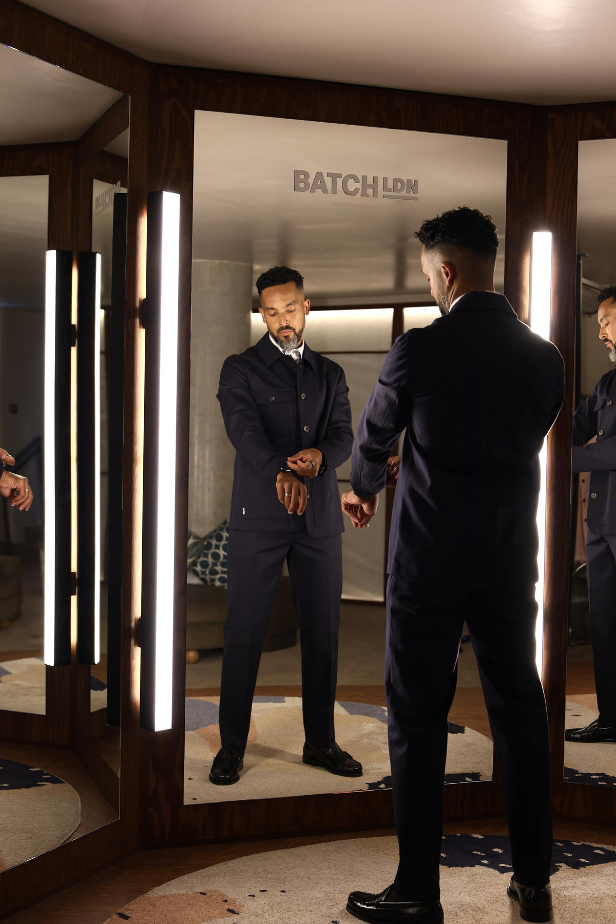 A man in a dark navy suit stands in front of a mirror in a modern fitting room, looking at his reflection. The room features wood paneling, vertical light strips, and a visible 'BATCH LDN' logo on the back of the mirror frame.