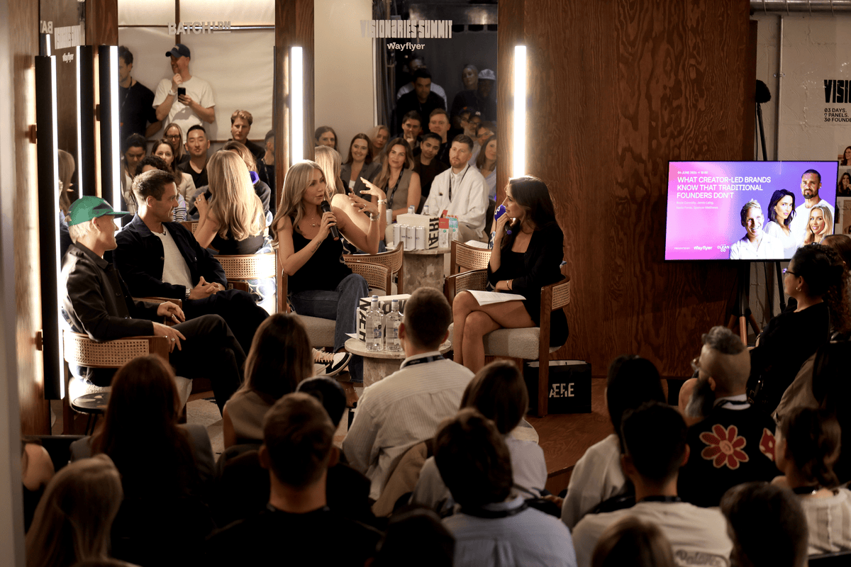 A panel discussion or talk show scene with multiple seated speakers and an audience in a studio or event space. A large screen on the right displays a colorful portrait graphic.